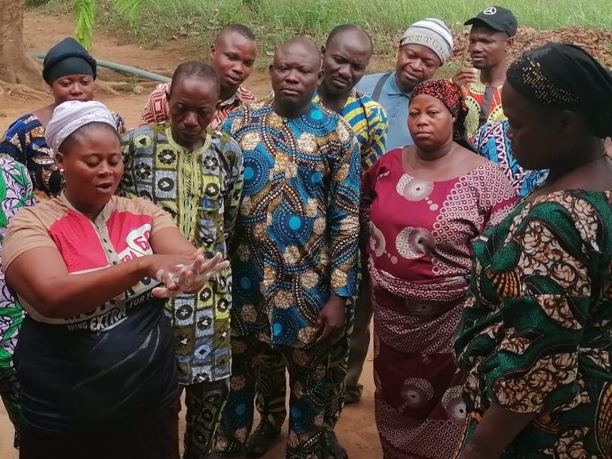 Joukko värikkäisiin afrikkalaisiin vaatteisiin pukeutuneita naisia ja miehiä seuraa, kun keskellä seisova nainen esittelee käsien pesua saippualla. Hänen edessään portailla on kaksi ämpäriä. Taustalla näkyy kaksi puuta ja niiden takana matalia rakennuksia.