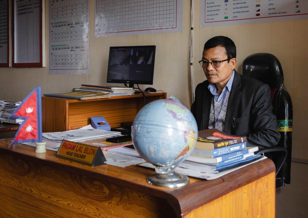 Keski-ikäinen silmälasipäinen nepalilainen mies istuu ison puisen työpöydän takana. Pöydällä on karttapallo, kirjapino, papereita ja pieni Nepalin lippu. Sivupöydällä on vihkoja ja tietokoneen näyttö. Takana seinällä on erilaisia taulukoita julistekoossa.