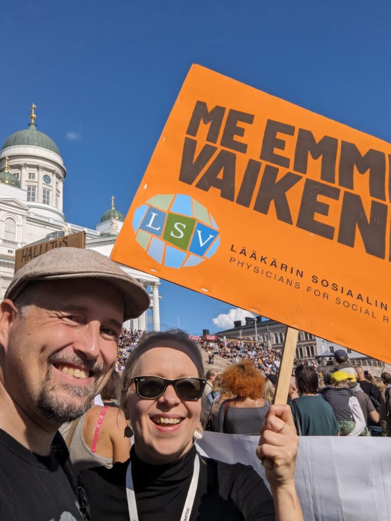 Mies ja nainen seisovat Helsingin Senaatintorilla iloisesti hymyillen. Nainen pitelee mielenosoituskylttiä, jossa lukee "Me emme vaikene" ja sen alla LSV:n logo. Taustalla näkyy muita mielenosoittajia ja Tuomiokirkko.