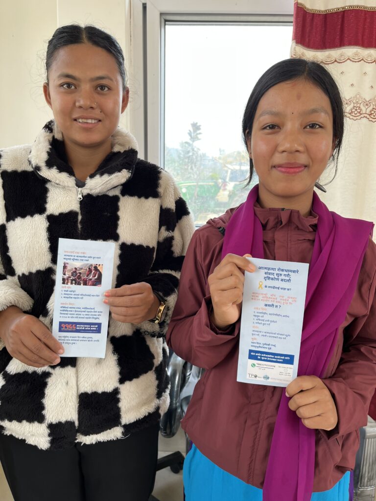 Kaksi nuorta nepalilaista naista seisoo vierekkäin ja katsoo kameraan hymyillen. Molemmilla on kädessään esite, jossa on nepalinkielistä tekstiä. Toinen on pukeutunut musta-valkoruudulliseen fleece-takkiin, toinen ohueen punaiseen takkiin ja aniliininpunaiseen huiviin. Taustalla näkyy toimiston ikkuna ja verho.