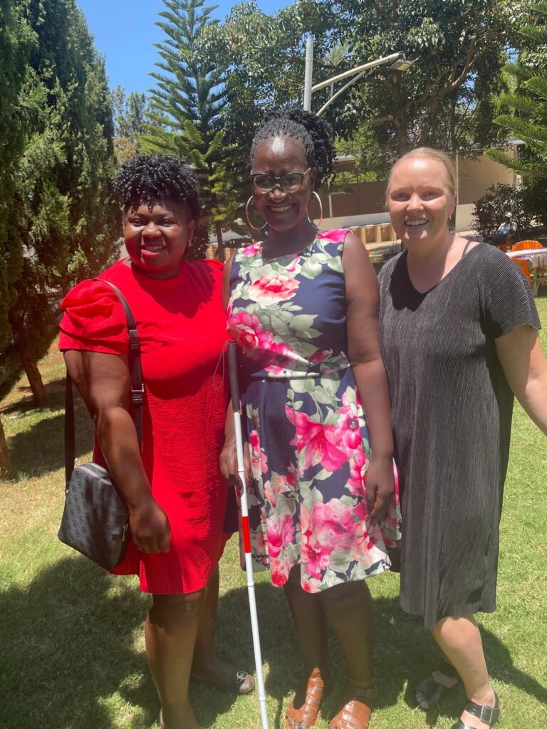 Three smiling women standing in a garden. The woman in the middle has a white cane in her hand.