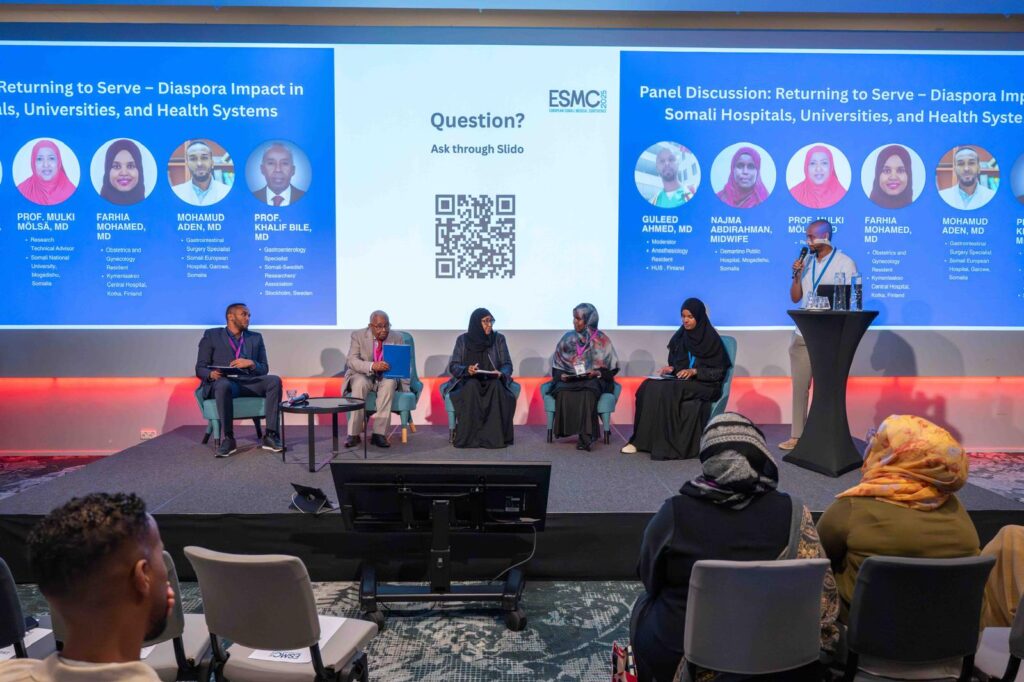 Luentosali, missä viisi panelistia istuu salin edessä ja moderaattori seisoo puhujapöntön takana ja puhuu mikrofoniin. Takaseinälle on heijastettu seminaarin ohjelma.