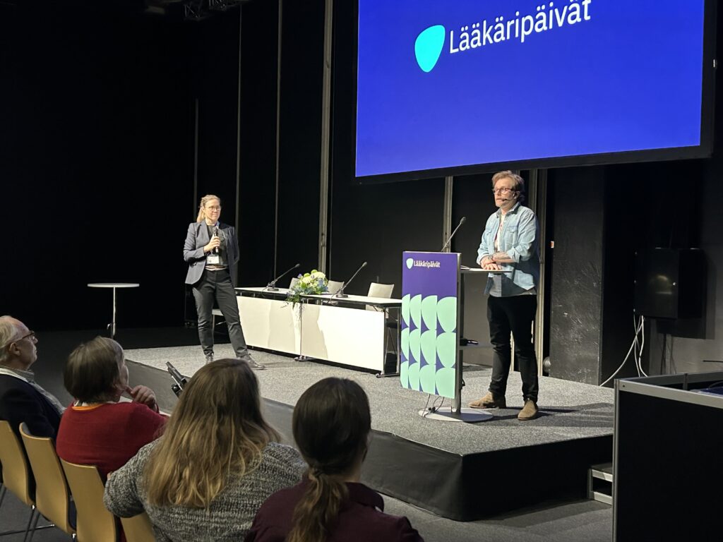 Puhuja ja juontaja seisovat luentosalin korokkeella. Etualallla näkyy eturivissä istuvia takaa ja taustaseinälle on heijastettu Lääkäripäivien nimi ja logo.