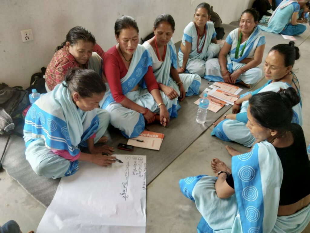 Ryhmä naisia samanlaisissa sinisissä sareissa istuu piirissä lattialla ja seuraavat kun yksi kirjoittaa isolle paperille nepalinkielistä tekstiä.